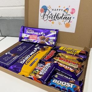 May include: A brown gift box containing a selection of Cadbury chocolate bars, such as Dairy Milk, Caramel, and Boost. A card with "Happy Birthday" is included, decorated with balloons and confetti. The box is filled with various chocolate bars.