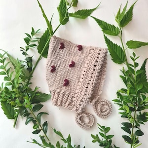 Può includere: Un berretto per neonato in maglia di colore rosa chiaro con un delicato bordo in pizzo e decorazioni a forma di cuore bordeaux.