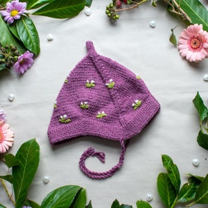 Könnte beinhalten: Eine lila gestrickte Baby-Mütze mit weißen Blumenstickereien. Die Mütze hat eine lange Schleife und ist von grünen Blättern und rosa Blüten umgeben.