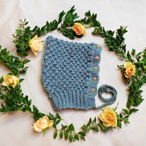 Puede incluir: Un gorro de bebé de ganchillo azul claro con bordado de flores rosas. El gorro está rodeado de una corona de hojas verdes y rosas amarillas.