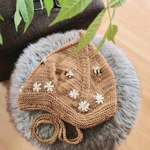 Peut inclure: Un bonnet en tricot marron avec un motif d'abeilles et de marguerites. Le bonnet a une longue ficelle qui peut être nouée sous le menton.