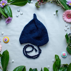 Peut inclure: Un bonnet bébé en tricot bleu marine avec un sommet pointu et une longue cravate. Le chapeau est entouré de feuilles vertes et de fleurs roses.
