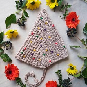 Könnte beinhalten: Eine beige gestrickte Mütze mit dreieckiger Form und einem langen Quasten. Die Mütze ist mit bunten Punkten verziert. Die Mütze ist von Blumen und Blättern umgeben.