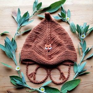 Puede incluir: Un gorro de bebé de punto marrón con un zorro bordado en la parte delantera. El gorro tiene una parte superior puntiaguda y lazos que se pueden atar debajo de la barbilla. El gorro está rodeado de hojas verdes de salvia.