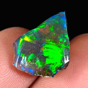 May include: A vibrant, raw opal gemstone with a dark base color. The opal displays a striking play-of-color, showcasing flashes of green, blue, and yellow. The gemstone is held between fingers against a black background.