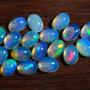 Può includere: Una collezione di opali ovali in varie tonalità di blu, con lampi iridescenti di verde, giallo e rosa. Le pietre preziose sono esposte su una superficie di legno marrone scuro, mostrando la loro bellezza naturale.