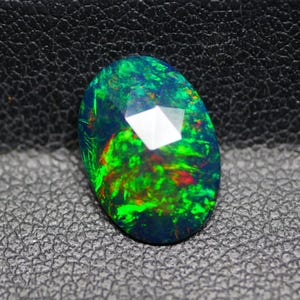 Puede incluir: Una gema ovalada con una superficie facetada. La piedra exhibe un llamativo juego de colores, con tonos verdes, rojos y azules dentro de una base oscura. La gema está sobre una superficie negra texturizada.
