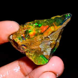 Puede incluir: Una gema de ópalo en bruto, sin cortar, con colores vibrantes. El ópalo muestra destellos de verde, naranja y azul dentro de una matriz de roca marrón y beige. La gema se sostiene en una mano, mostrando su belleza natural.