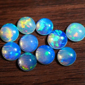 Può includere: Una collezione di dieci opali rotondi, ognuno con un vibrante gioco di colori. Gli opali variano nelle tonalità del blu, con lampi di verde, giallo e rosso. Le pietre preziose sono su una superficie di legno scuro.
