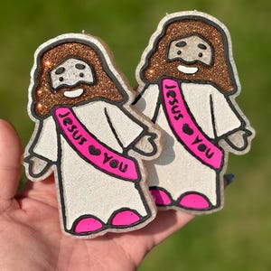 May include: Two glittery Jesus-shaped ornaments. Each has a brown glitter halo and beard, a white robe, and a pink sash that reads "Jesus You." The ornaments have black outlines and pink accents.