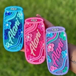 May include: Three colorful, glittery can-shaped accessories. One is blue, one is pink, and one is purple. Each has the name "Alani" in cursive and floral accents. The background is a blurred green.