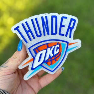 May include: A clear, glittery Oklahoma City Thunder logo with blue lettering and accents. The logo features the word "THUNDER" at the top and "OKC" inside a shield with an orange basketball design. The background is a blurred green.