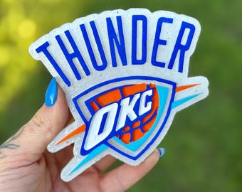 May include: A clear, glittery Oklahoma City Thunder logo with blue lettering and accents. The logo features the word "THUNDER" at the top and "OKC" inside a shield with an orange basketball design. The background is a blurred green.