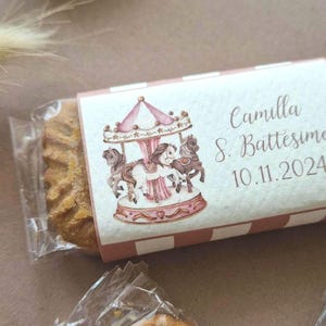 Può includere: Una scatola regalo rosa e bianca con un'illustrazione di giostra e il testo "Camilla S. Battesimo 10.11.2024". La scatola è avvolta in cellophane trasparente e contiene un biscotto.