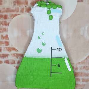 May include: A green and white science beaker piñata with a blue and white measuring scale and the numbers 5 and 10. The piñata is filled with green confetti.
