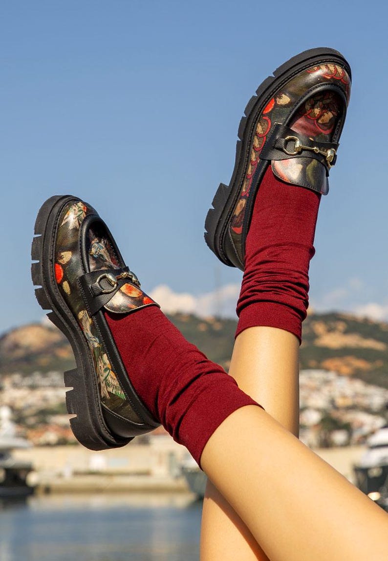 May include: A pair of black loafers with a multi-colored floral and bird print, featuring a gold-tone metal horsebit detail across the vamp and a thick, lugged black rubber sole. The loafers are worn with solid dark burgundy crew-length socks.