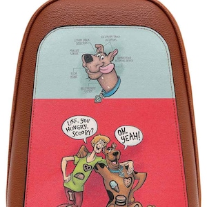 Puede incluir: Una mochila de piel sintética marrón con un gráfico de Scooby-Doo. El panel superior muestra un diagrama de Scooby-Doo con etiquetas como "Scooby Snack Detectors". El panel inferior muestra a Shaggy y Scooby-Doo con bocadillos que dicen "Like, you hungry, Scooby?" y "Oh, yeah!"