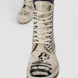 May include: Beige lace-up boots featuring a black and white striped pattern on the tongue and sides. The toe area displays a portrait of Charlie Chaplin in a striped prison uniform. The text A DAY WITHOUT LAUGHTER IS A DAY WASTED is printed.