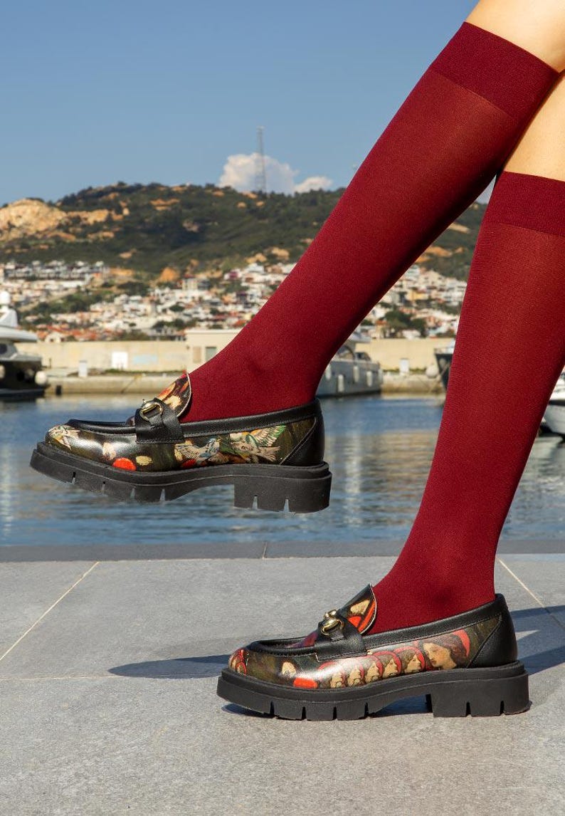 May include: A pair of black loafers featuring a multi-colored graphic print of classical figures and birds, accented with a gold-tone metal horsebit detail on the vamp. The shoes have a thick, black lug sole and are worn with solid dark red knee-high socks.