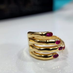 Op de afbeelding: Een goudkleurige ring met een uniek ontwerp. De ring heeft een reeks gebogen, vingerachtige vormen, elk met een kleine, ovale rode edelsteen. De ring ligt op een witte ondergrond.