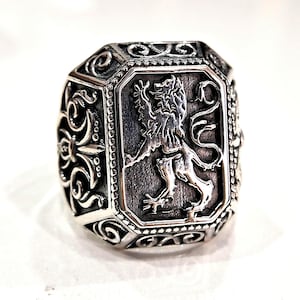 Può includere: Un anello color argento con una faccia ottagonale con un emblema di leone dettagliato. L'anello presenta volute ornate e una finitura strutturata, che mostra un classico design araldico. L'anello è un pezzo importante.