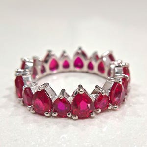 Op de afbeelding: Een zilveren ring met een doorlopende rij levendige, druppelvormige, rode edelstenen. De edelstenen zijn dicht op elkaar gezet, waardoor een volledige band van kleur ontstaat. De ring wordt gepresenteerd tegen een witte achtergrond, wat het ontwerp van de sieraden benadrukt.