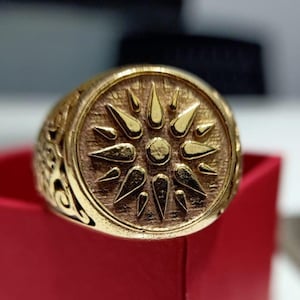 May include: A gold-colored ring with a sunburst design on the face. The ring has a detailed, textured surface and a thick band. The ring is set against a red background, suggesting a jewelry display.