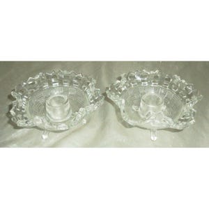 Könnte beinhalten: Zwei Kerzenhalter aus klarem Glas. Jeder hat eine Schale mit Korbwebmuster, einem gerüschten Rand und einer zentralen Kerzenschale. Die Kerzenhalter werden von drei kleinen Füßen getragen.