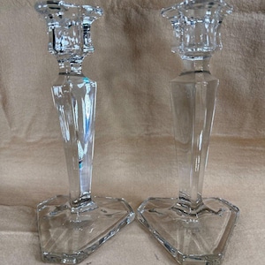 Puede incluir: Dos candelabros de cristal transparente. Cada uno tiene un diseño geométrico facetado con una parte superior cuadrada y una base triangular. Los candelabros son transparentes y reflejan la luz.