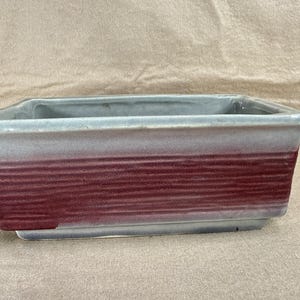 Puede incluir: Jardinera rectangular de cerámica con diseño bicolor. La parte superior es gris claro, que transiciona a una banda roja oscura en el medio. La jardinera tiene una forma simple y moderna, adecuada para varias plantas.