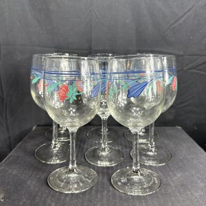 May include: A set of six clear glass wine glasses, each featuring a delicate painted floral motif around the upper bowl. The design includes stylised red and blue flowers with green leaves, accented by thin horizontal blue lines encircling the glass.
