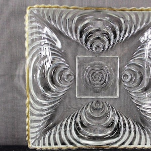 May include: Clear glass square dish with a gold rim. The dish features a central square with a spiral design, surrounded by four spiral designs. The edges of the dish have a wavy pattern.