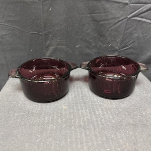 May include: Two matching, dark purple glass bowls with handles. The bowls are round with a slightly flared rim and are made of a transparent glass. The bowls are sitting on a grey surface.