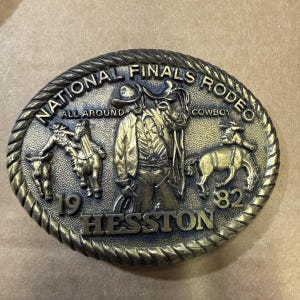 Könnte beinhalten: Eine ovale Gürtelschnalle in Messingoptik mit einem Reliefdesign eines Cowboys, eines Pferdes und eines Stiers. Der Text lautet NATIONAL FINALS RODEO, ALL AROUND COWBOY, 1982 und HESSTON. Der Rand hat eine gedrehte Seilstruktur.