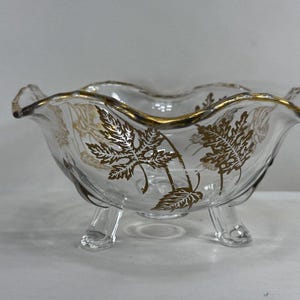 May include: Clear glass footed bowl with a scalloped rim and gold trim. The bowl features a gold leaf pattern and is supported by three clear glass feet. A decorative piece for serving or display.
