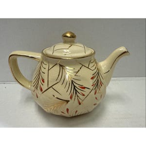 Può includere: Teiera color crema con manico, beccuccio e coperchio dorati. La teiera è decorata con motivi dorati e rossi a forma di foglie. Il coperchio ha un pomello dorato.