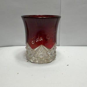 May include: A deep red glass tumbler featuring a clear, faceted base with a starburst pattern. The upper portion of the glass is smooth and translucent red, with the cursive script "Ida" etched into the surface. The vessel has a wide, flared rim.