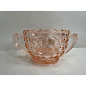 May include: A pink glass sugar bowl with two handles. The bowl features a geometric, diamond-like pattern and a scalloped rim. The sugar bowl is a light pink colour and sits on a small, round base.