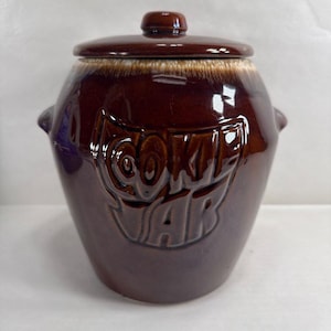 Puede incluir: Un tarro de galletas de cerámica marrón con tapa. El tarro tiene forma redondeada con dos asas pequeñas y las palabras "COOKIE JAR" en relieve en la parte delantera. La tapa también es marrón y tiene un pequeño pomo.