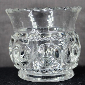 Puede incluir: Portavelas votivo de cristal transparente con borde festoneado y un patrón en relieve detallado de círculos y diseños florales. La base es un pequeño pedestal circular. El cristal es transparente.