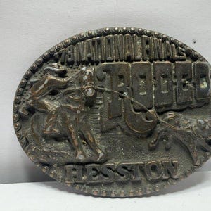 Op de afbeelding: Een ovale riemgesp van bronskleurig metaal met een reliëf van een persoon op een bokkend paard en een kalf. De tekst luidt 78 NATIONAL FINALS RODEO en HESSTON. De rand is voorzien van een kralenstructuur rondom de gehele omtrek.