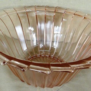 May include: A vintage, light pink glass bowl with a scalloped rim and vertical ribbed design. The bowl has a rounded shape and is likely used for serving or display. The glass appears to be smooth and transparent.