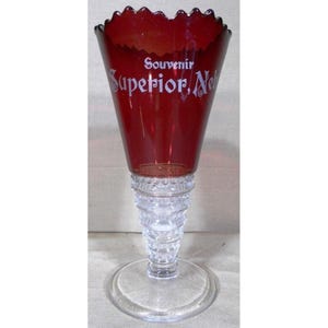 以下が含まれることがあります： ルビーレッドのフレアボウルと透明で質感のあるステムとベースを備えた、ヴィンテージのお土産グラス。グラスには「Souvenir Superior, Neb」の文字が白で書かれています。ボウルの縁はスカラップエッジです。