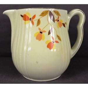 May include: Cream-colored ceramic pitcher with a floral design. The pitcher features a curved handle and a spout. The floral design includes orange, yellow, and brown flowers and leaves. The rim and handle are trimmed with gold.
