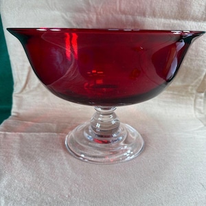 May include: A ruby red glass bowl with a clear glass pedestal base. The bowl has a wide, flared rim and a rounded body. The pedestal base is circular and has a ribbed design. The bowl is likely used for serving or display.