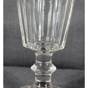 May include: Clear, vintage-style stemmed glass with a fluted bowl. The glass has a wide, slightly flared rim and a detailed stem, resting on a circular base. The glass is likely used for serving beverages.