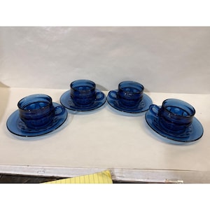 May include: Four cobalt blue glass teacups and saucers. Each cup and saucer has a matching embossed pattern. The cups have handles and sit on oval-shaped saucers. The set is arranged on a white surface.