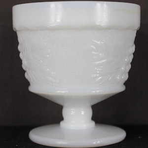 May include: A white, vintage-style glass goblet with a textured, floral design. The goblet has a wide rim, a textured body, a stem, and a circular base. The glass has a milky, opaque appearance, and the goblet is likely made of milk glass.