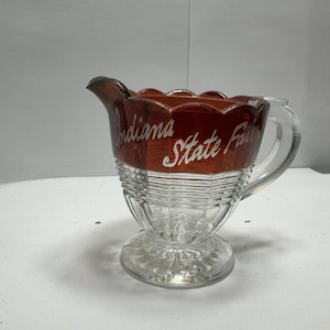 May include: A clear glass creamer pitcher featuring a scalloped rim and a pedestal base. The upper portion of the pitcher is amber-colored glass with the white script text Indiana State Fair printed on the side. It includes a handle and a spout.