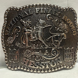 以下が含まれることがあります： 銀色の金属製ベルトバックル。質感のある背景に「NATIONAL FINALS RODEO」と「HESSTON」の浮き彫り文字が施されています。中央には馬に乗ったカウボーイが子牛に投げ縄をかける様子が描かれ、両脇には「NFR 19」と「NFR 85」と記された2つの盾が配置されています。
